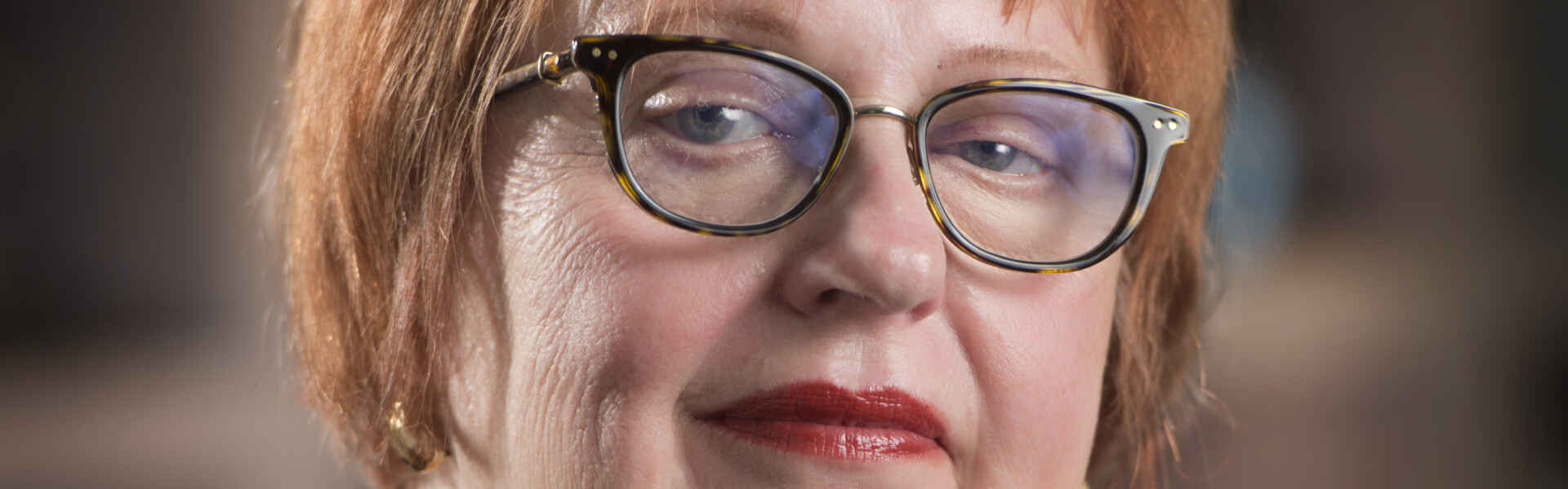 a person with short red hair and glasses wearing a brown jacket poses for a headshot