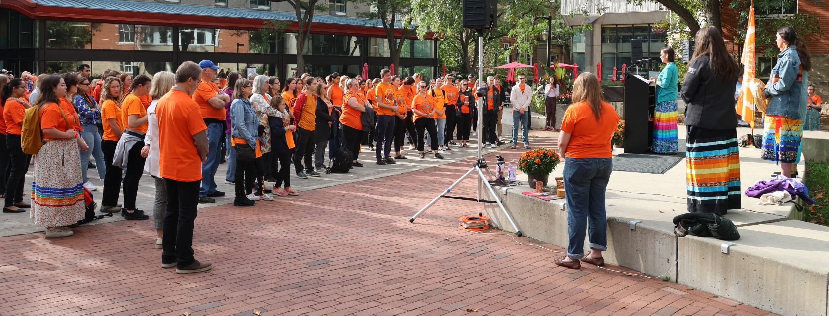 A group of people gather in Branion Plaza wearing Orange Shirts and listen to a speaker at a podium.