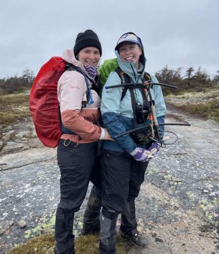 A person in an orange coat, black toque, black pants and red backpack stands smiling at the camera beside a person in a green and blue jacket, baseball cap and black pants smiling at the camera and holding equipment to study a herd of caribou in Newfoundland.