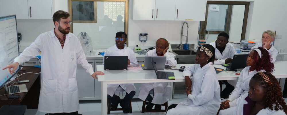 A man with a beard and glasses wearing a white lab coat stands pointing at a reference in front of students in white lab coats seated at desks with laptops in front of them.