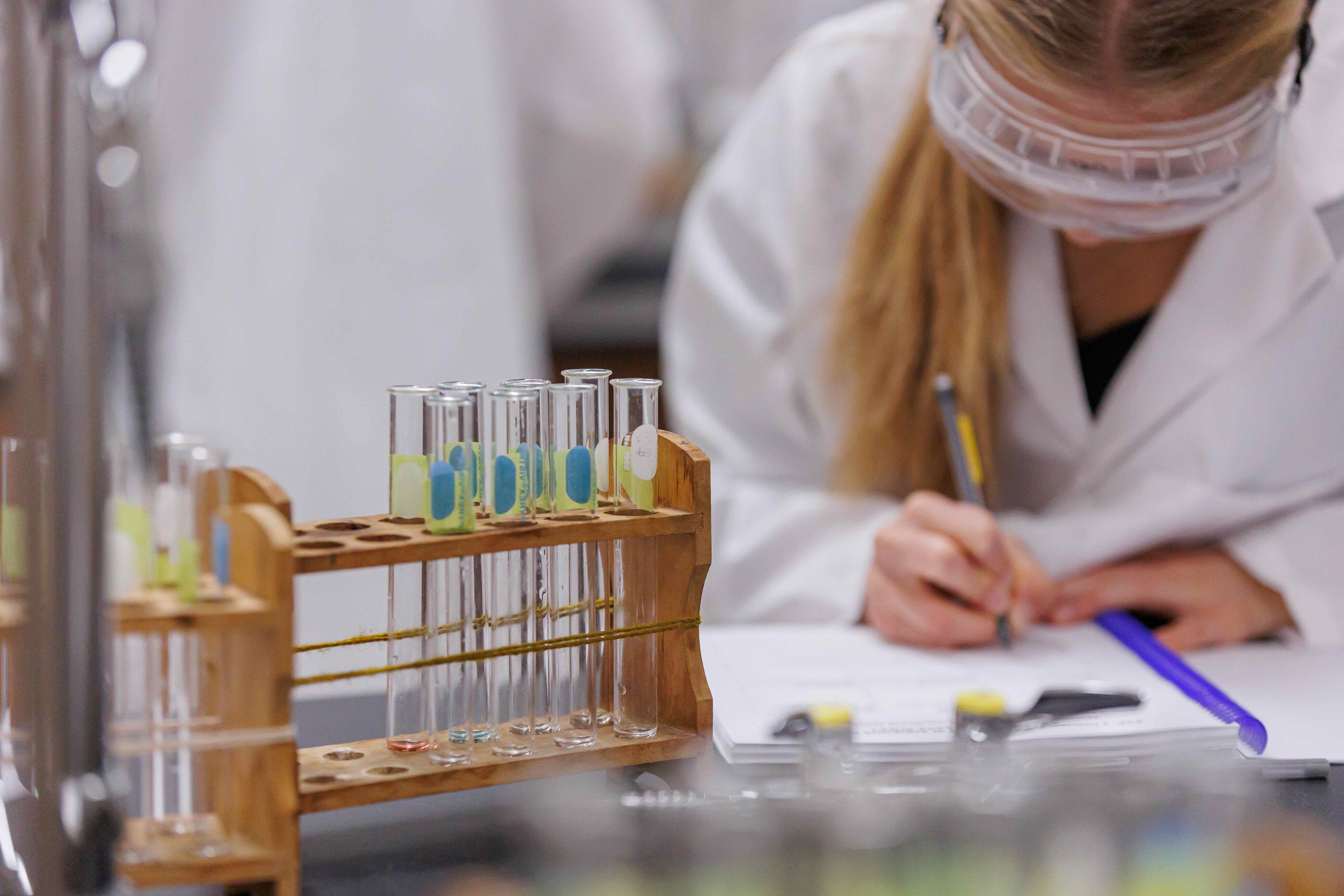 A student in a white lab coat and safety goggles is taking notes in a chemistry lab. In the foreground, a wooden test tube rack holds multiple labeled test tubes, some with small amounts of liquid in light green, blue, and orange tones. The focus is on the equipment and the student's hand as she writes in a notebook, with the background slightly blurred to emphasize the experimental setup.