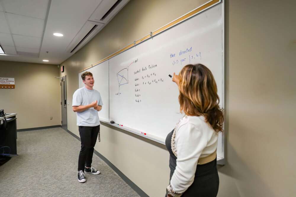 A student points to a whiteboard while explaining the “Shortest Route Problem” in a math classroom. Another student listens with interest. The whiteboard includes a diagram and formulas, with notes such as “Blue: directed” and “O-D pair: {2, 4}”.