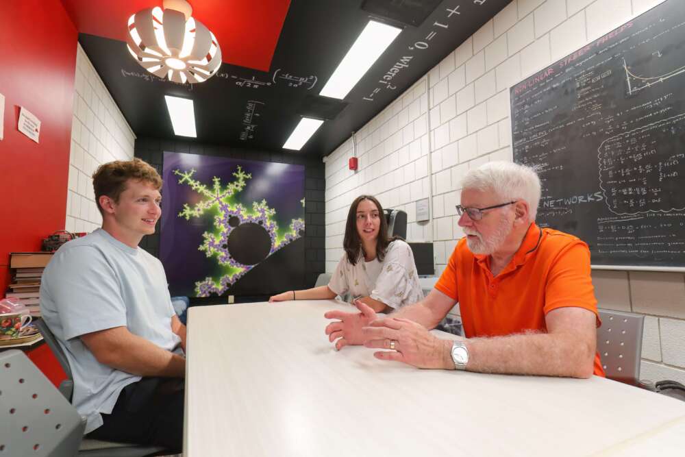 Three people sit at a table in a colourful math lounge featuring a fractal mural and ceiling equations. Math professor Gary Umphrey (in orange) speaks to two graduate students. Behind them are chalkboards filled with equations and a title reading “Non-Linear Street Graphs”.