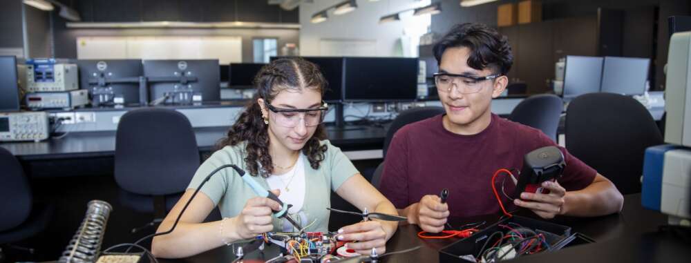 Two students wearing safety glasses work together on drone components in an engineering lab. One student uses a soldering iron while the other holds a controller with red wires. Computer monitors and testing equipment line the benches behind them.