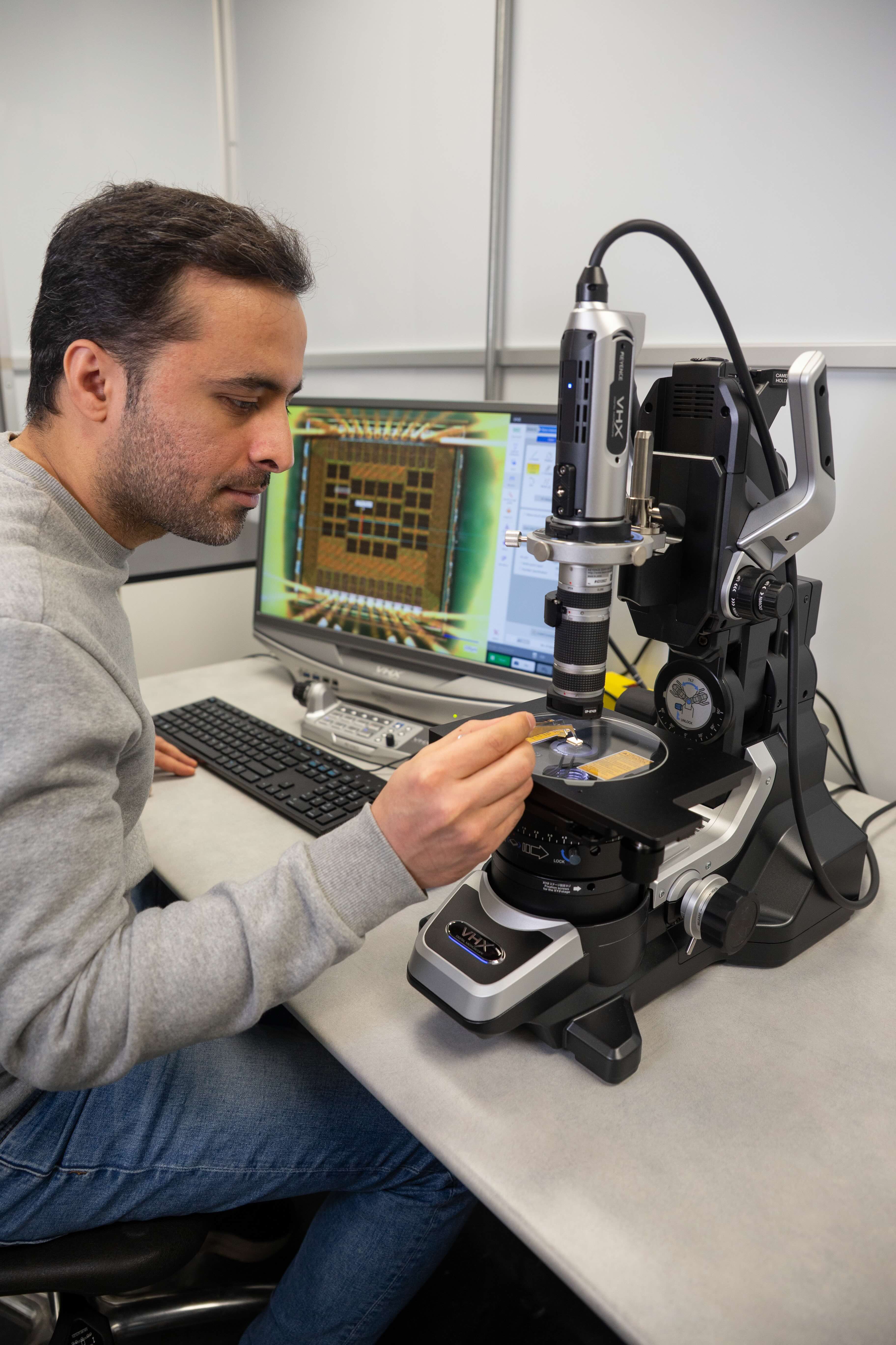 A researcher uses a precision microscope to examine a computer chip, carefully holding a probe tool. The computer monitor in the background displays a magnified image of the chip.