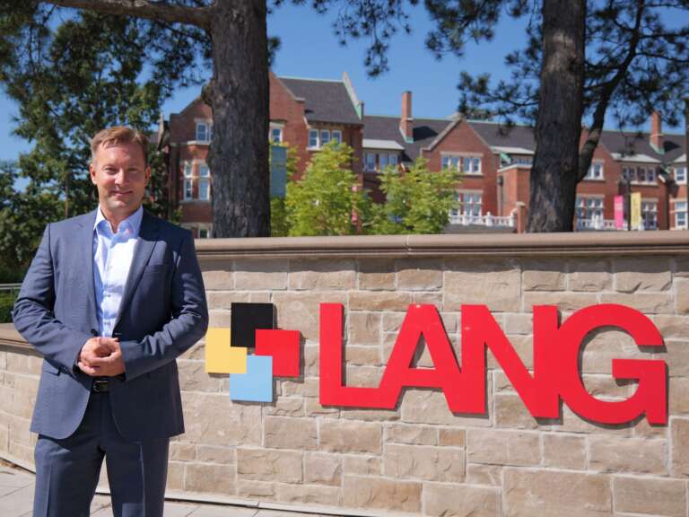 a person in a blue suit and white shirt poses in front of a beige brick wall with a sign that says lang in red letters