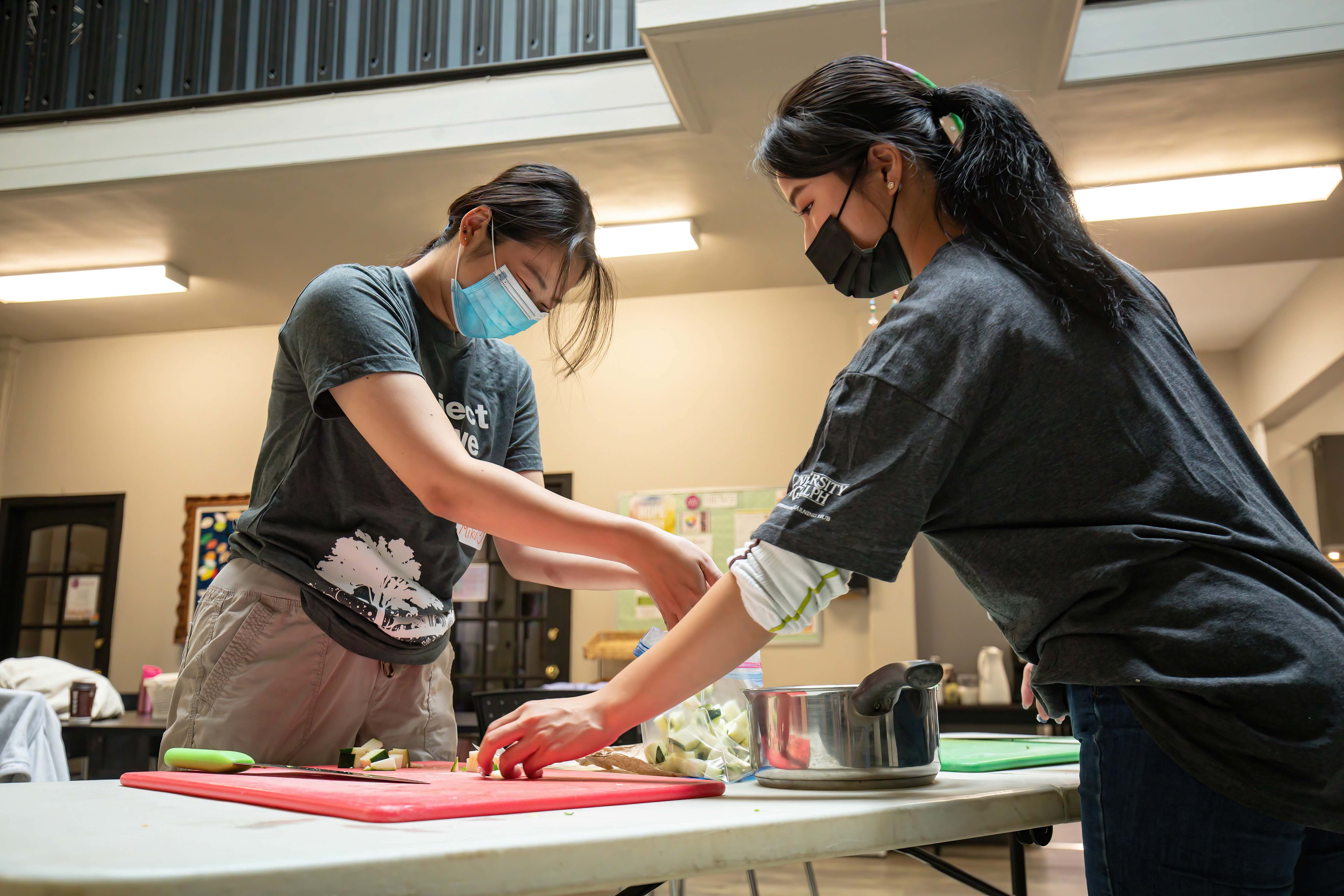 Two students wearing charcoal grey “Project Serve” T-shirts and medical masks prepare food together indoors. One is slicing vegetables on a red cutting board while the other assists. A pot and a bag of cubed vegetables sit on the table in front of them. Bulletin boards and a soft-lit interior are visible in the background.