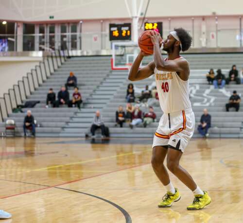 A man in a white Gryphon basketball uniform and yellow gym shoes stands poised to throw a basketball while spectators on bleachers watch in the background.