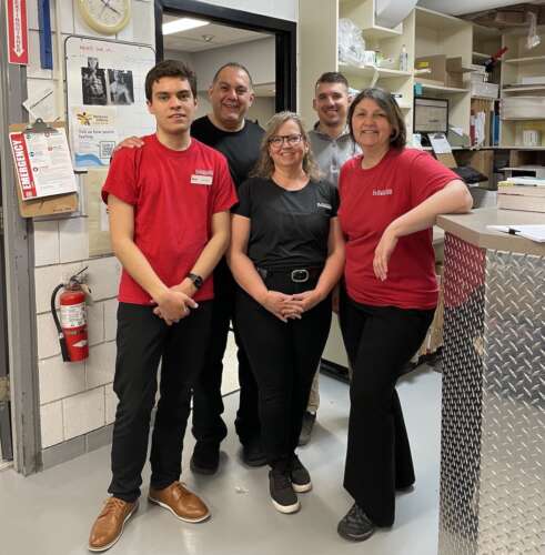 A group of five people stand together in red and black t-shirts in the storage room of the University Bookstore.