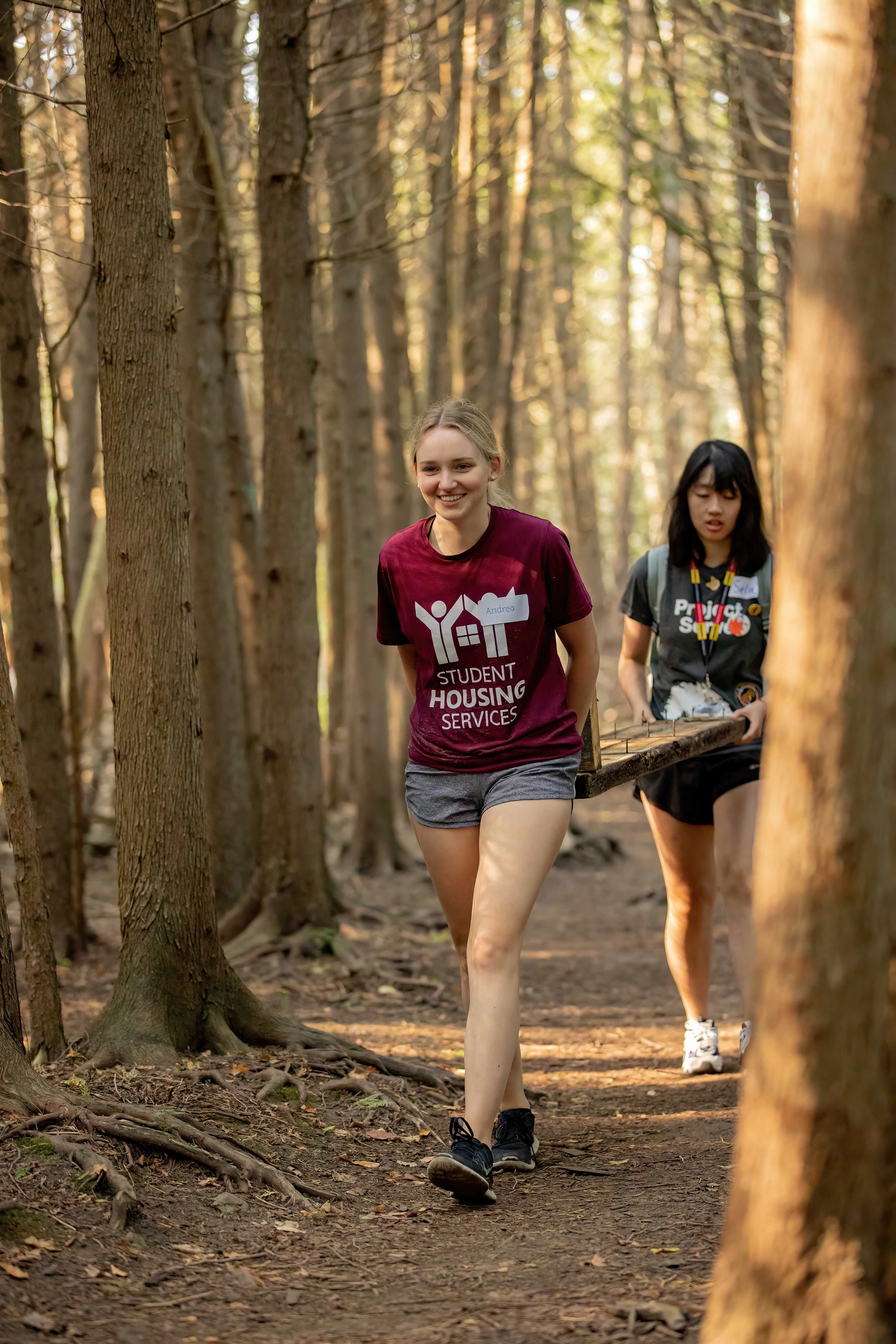 Two students walk along a wooded trail, carrying a wooden plank together. The student in front wears a burgundy “Student Housing Services” T-shirt and shorts, smiling as she walks. Her name tag reads “Andrea.” Behind her, another student wears a dark grey “Project Serve” T-shirt and shorts. Tall trees surround them in every direction, casting dappled light on the forest path.