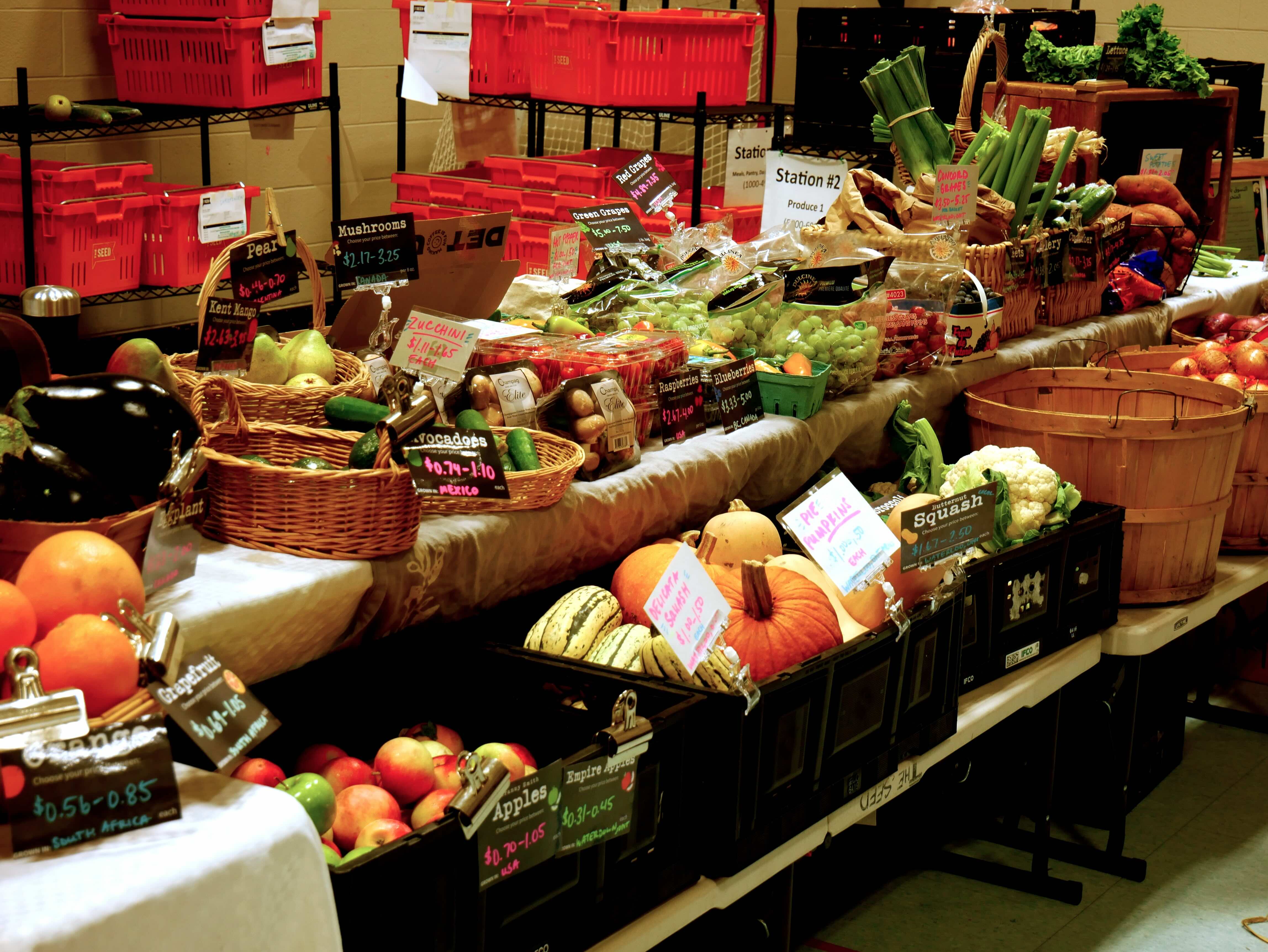 A long table lined with fresh produce—pumpkins, apples, squash, grapes, avocados, cauliflower, and more—sits inside a community centre. Handmade signs list prices and origins, such as “Avocados – Mexico – $0.71–1.10” and “Zucchini Singles – $0.87.” In the background are bright red storage bins and signage for Station #2.