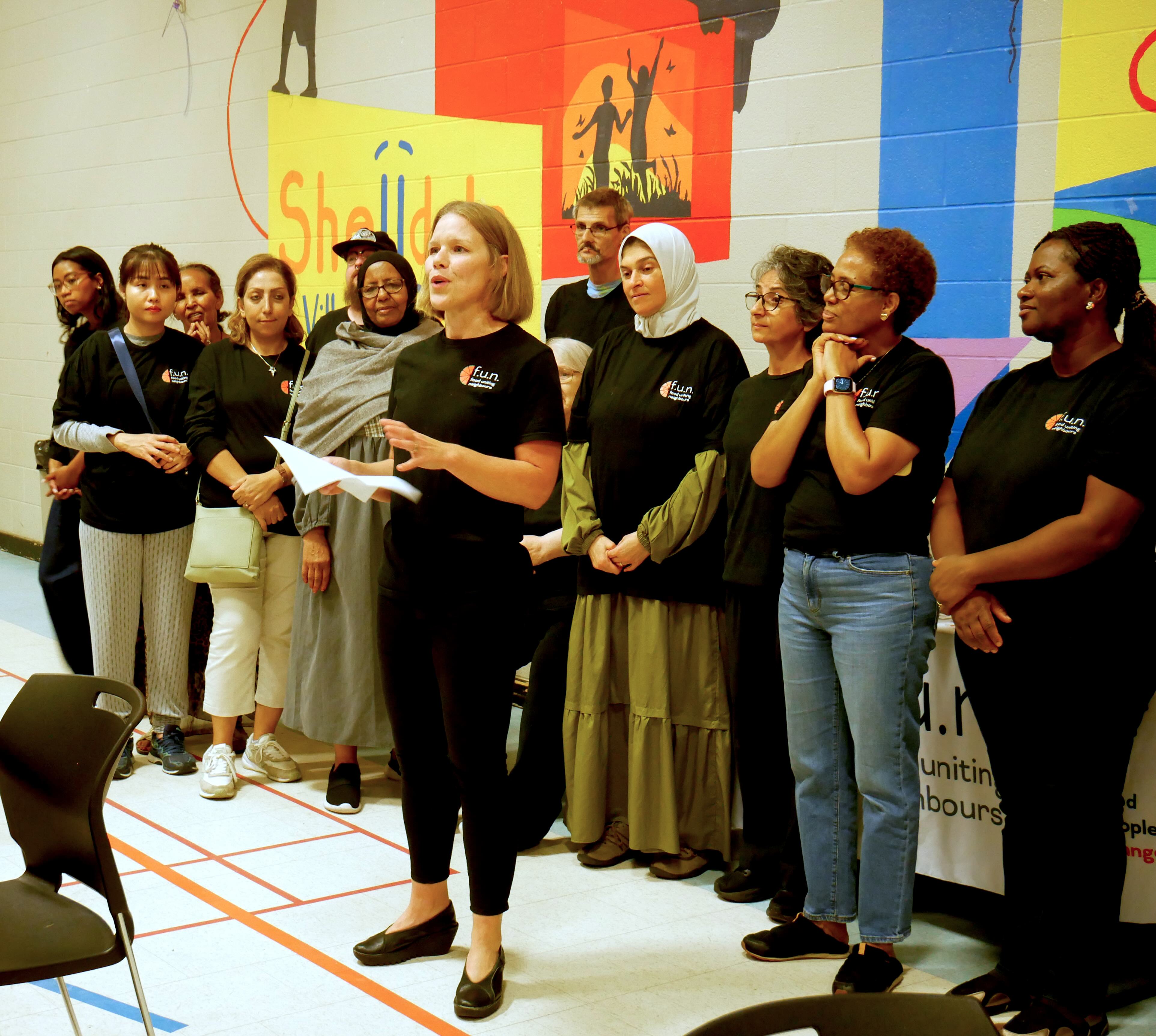 A group of diverse women and men stand in a line inside a community gym, all wearing matching “fun” logo t-shirts. Dr. Jess Haines in the centre speaks while holding a sheet of paper, as others listen or smile. Behind them, a mural reads “Shelldale” in bright yellow and red.