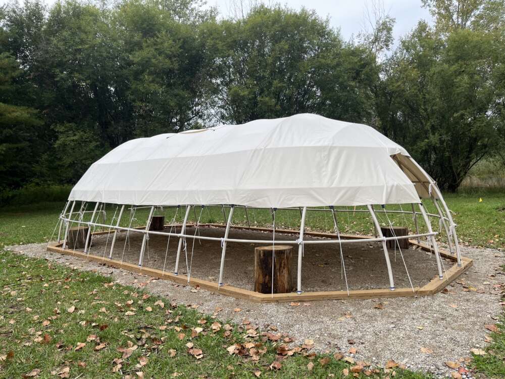a structure made of white pvc pipe with a beige canvas covering, with tree trunks along the edges, sits in a patch gravel surrounded by green grass and trees
