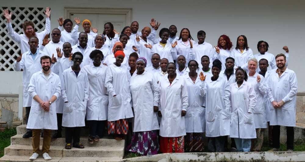 A group of scientists stand in three rows together wearing lab coats over their multi-coloured clothing.