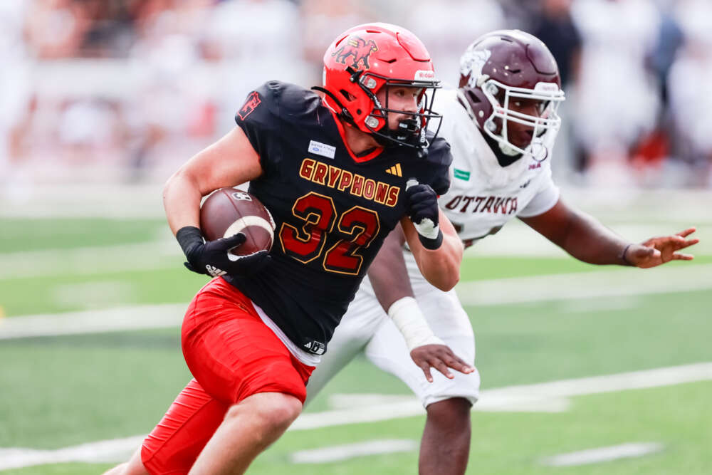 a football player in black and red with gryphons and 32 on their jersey holding a brown ball runs in front of a second player dressed in white