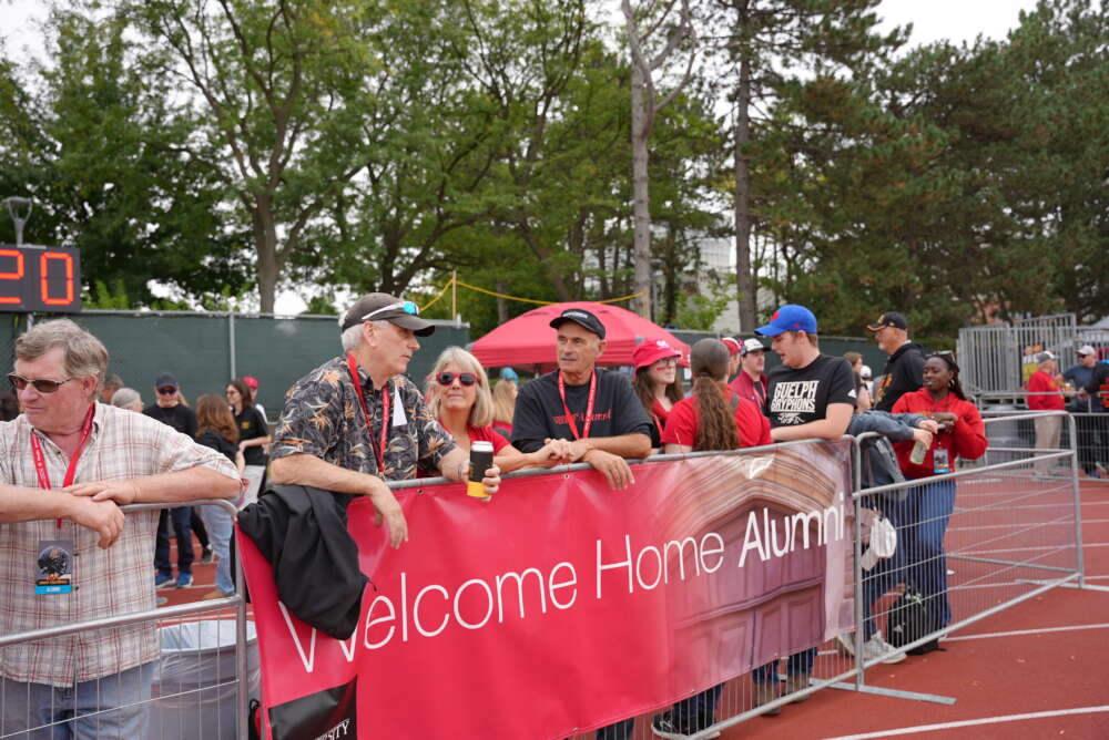 fans stand behind a barrier with a red sign that says welcome home alumni 