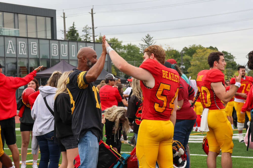 a football player in a red jersey and yellow pants high fives a fan in a black jersey and blue jeans on a football field