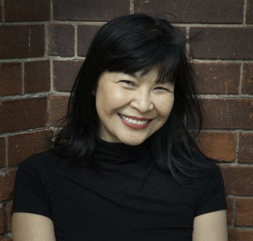 A person with shoulder length black hair and bangs stands smiling into the camera wearing a black turtleneck t-shirt in front of a red brick wall.