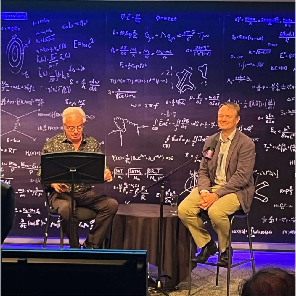 two people sit on tall stools on a stage with a black background with white writing on it