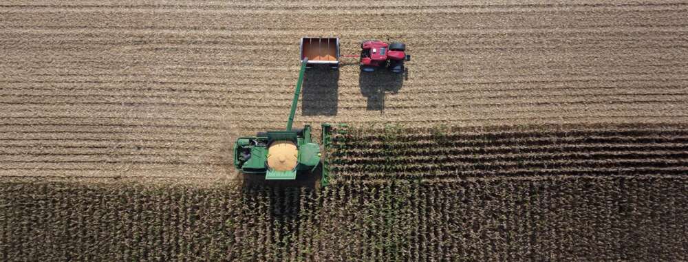 An aerial view of a combine harvester transferring corn into a red tractor trailer in a brown-striped field. The image illustrates the mechanized precision of agricultural harvesting