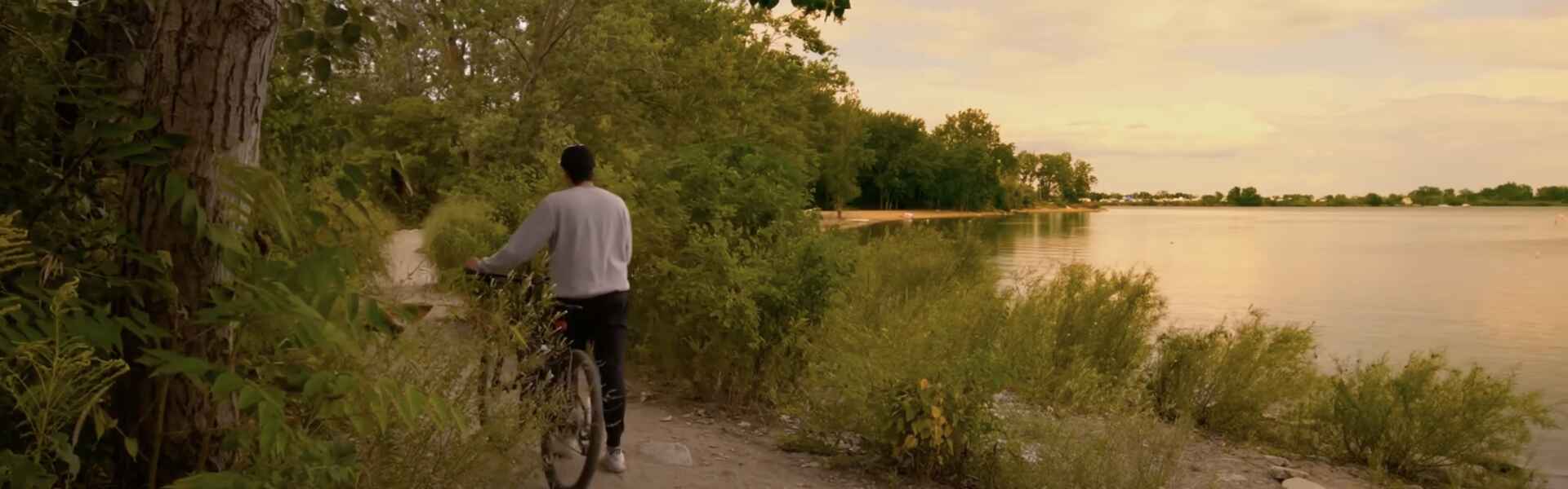 A person wearing a grey sweatshirt and black åsweatpants walks a black bicycle down a gravel path through greenery beside a lake as the sun sets in a pink sky.