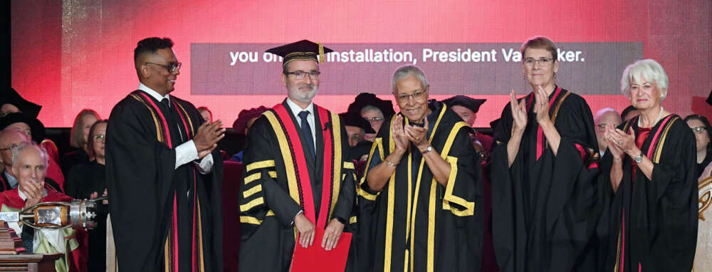 Four people wearing robes on a stage applaud while a man in regalia robes and cap smiles and holds a red folder
