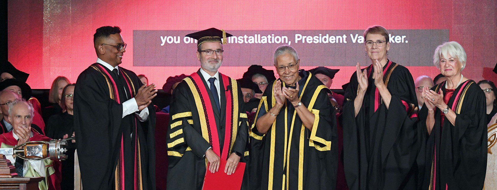 Four people wearing robes on a stage applaud while a man in regalia robes and cap smiles and holds a red folder