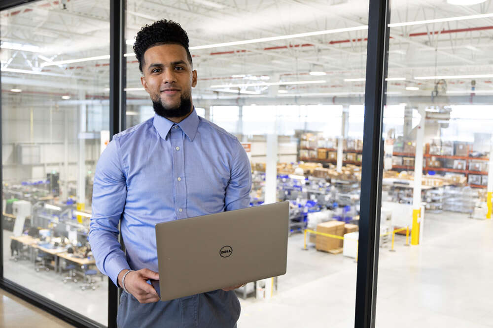 A co-op student holds an open laptop standing before windows looking onto a factory floor