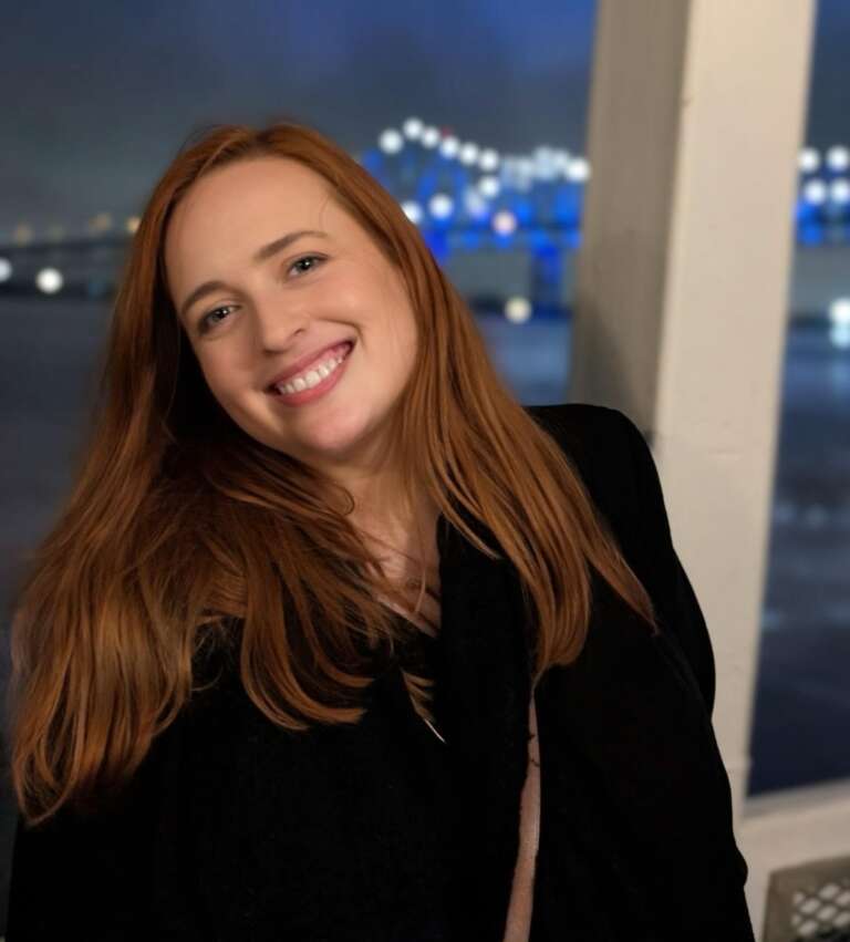 A person with long red hair wearing a black coat with a brown leather strap bag across their chest stands smiling into the camera on a ferry in water with a bridge lit with blue lights in the background.