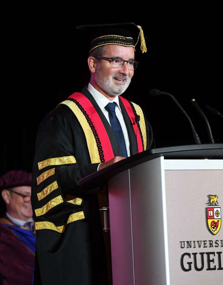 A man wearing regalia robes and cap smiles while standing at a University of Guelph podium