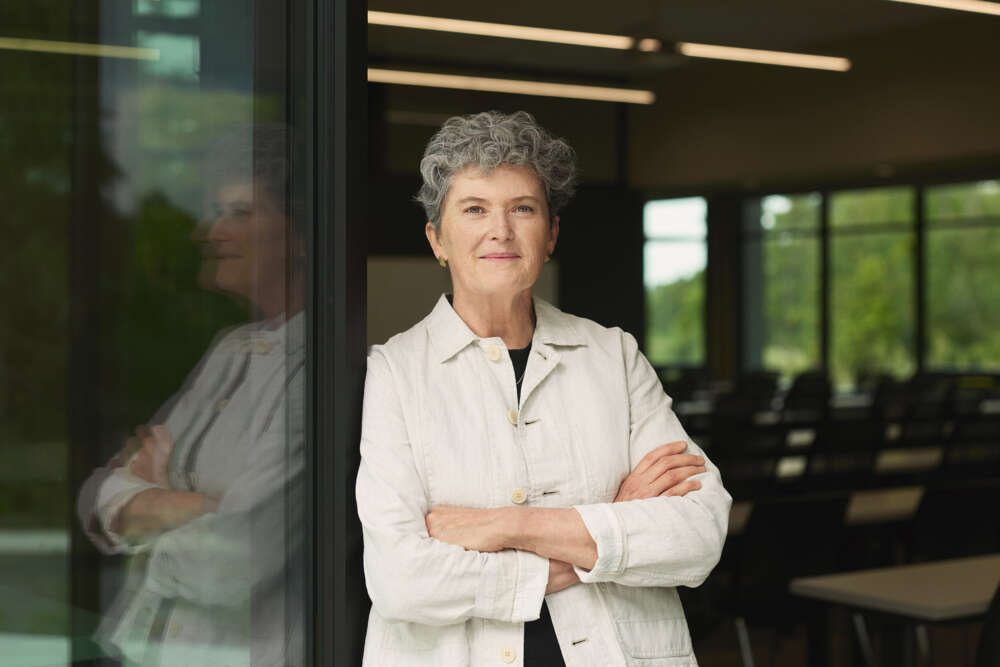 a person with short grey hair poses for a portrait in front of a room filled with rows of tables and chairs