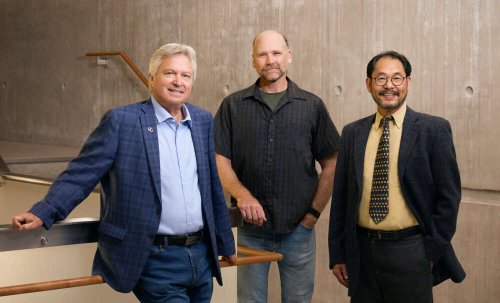 Three men smile and pose for a portrait while standing near stairs