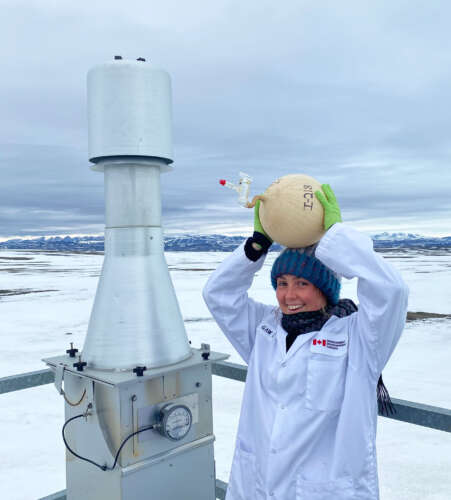 A person in a white lab coat and blue toque and green gloves stands beside equipment in the Arctic where the ground is covered in snow.