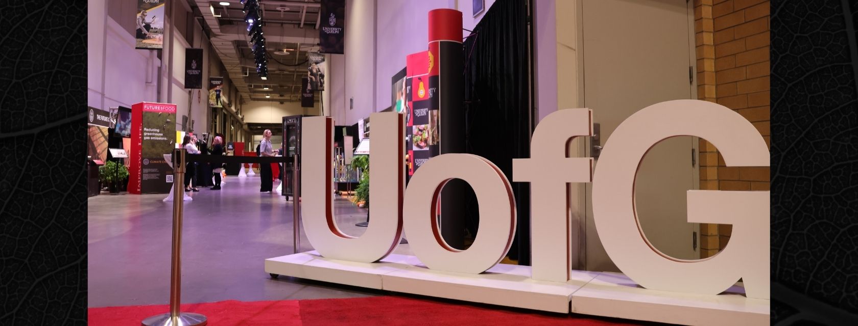 A hallway at the Royal Agricultural Winter Fair is decorated with banners and exhibits promoting the University of Guelph. In the foreground, large white 3D letters spell “U of G” atop a red carpet. Visitors browse displays in the background under purple spotlights.
