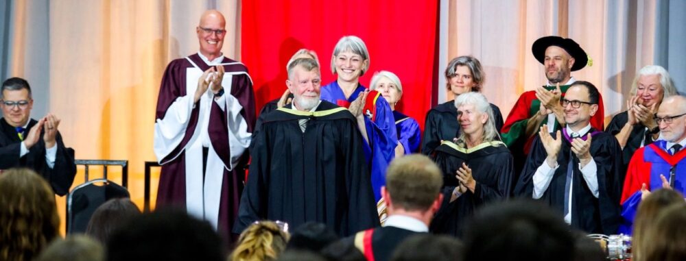 A man with a beard and gray hair wearing a black graduation gown stands on a convocation stage to receive his degree with a group of University leadership standing behind him and an audience in front of them.