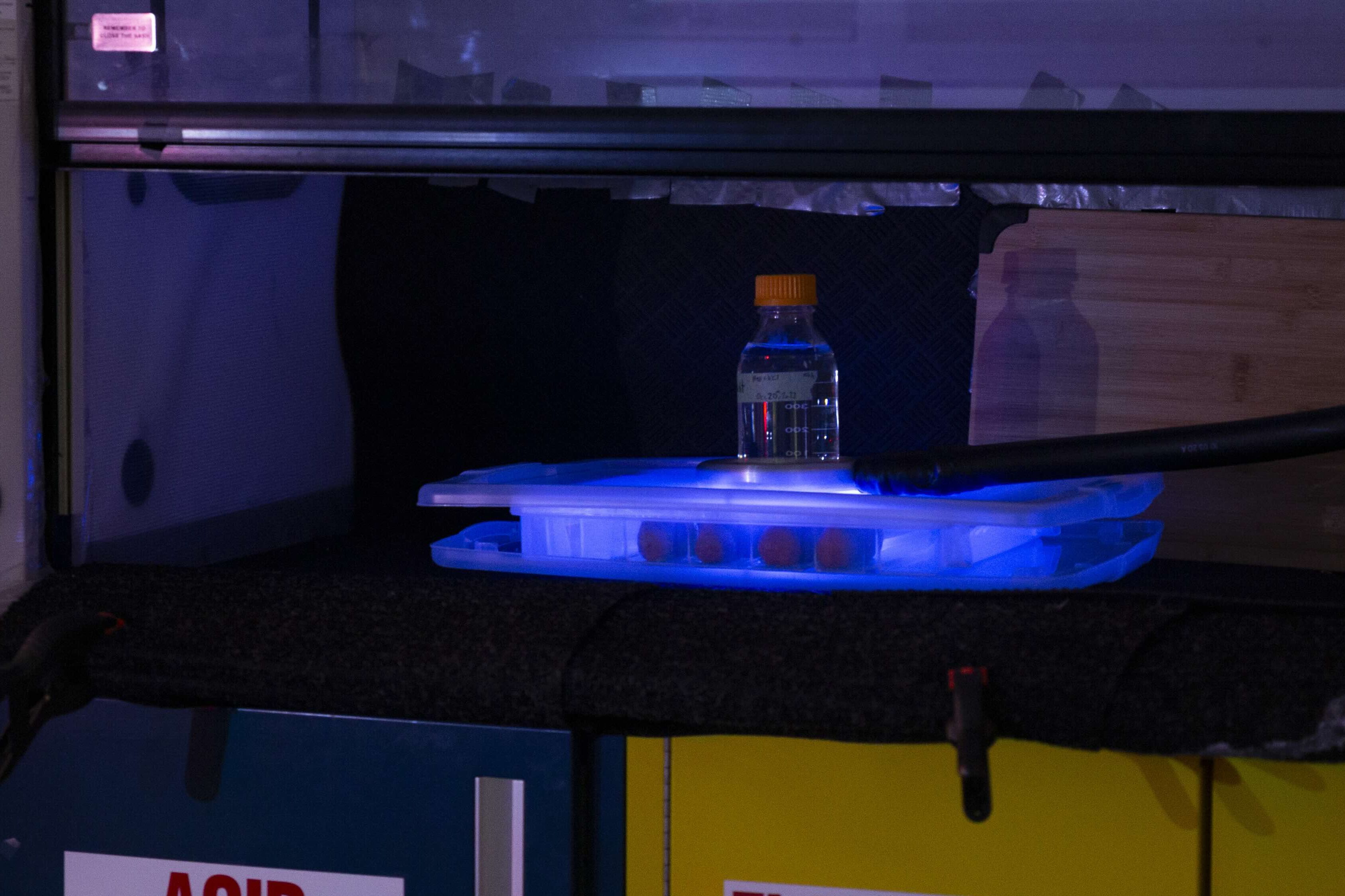 Lab sample glows blue under fume hood with safety signs below.