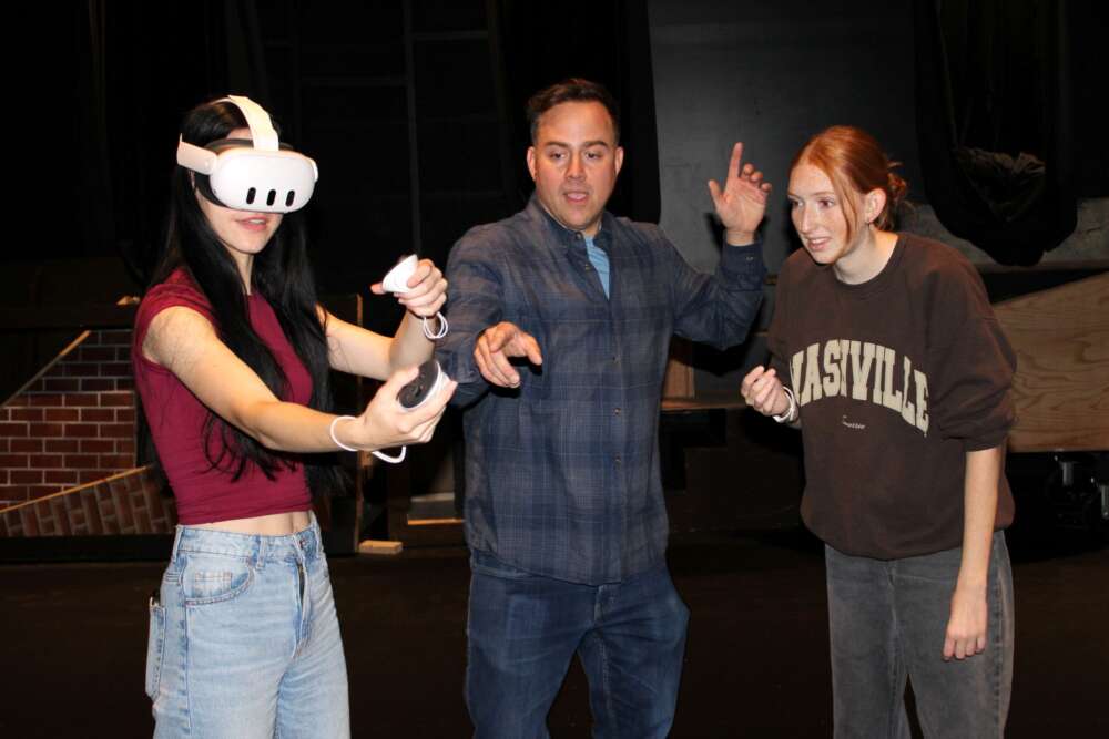 A student wearing a VR headset in a theatre lab is directed by a man on how to use the handset while another student observes