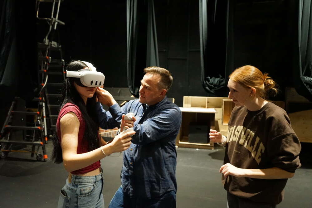 Three people stand in a theatre set with black velvet curtains behind them. One person adjusts a VR headset onto the head of another