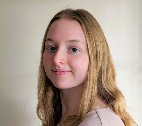 A person with long blonde hair sits on an angle looking into the camera in front of a beige background.