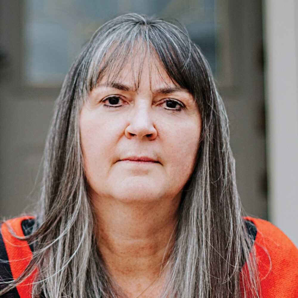 A person with long, gray and brown hair with bangs wears a red sweater and looks into the camera in front of a gray stone building.