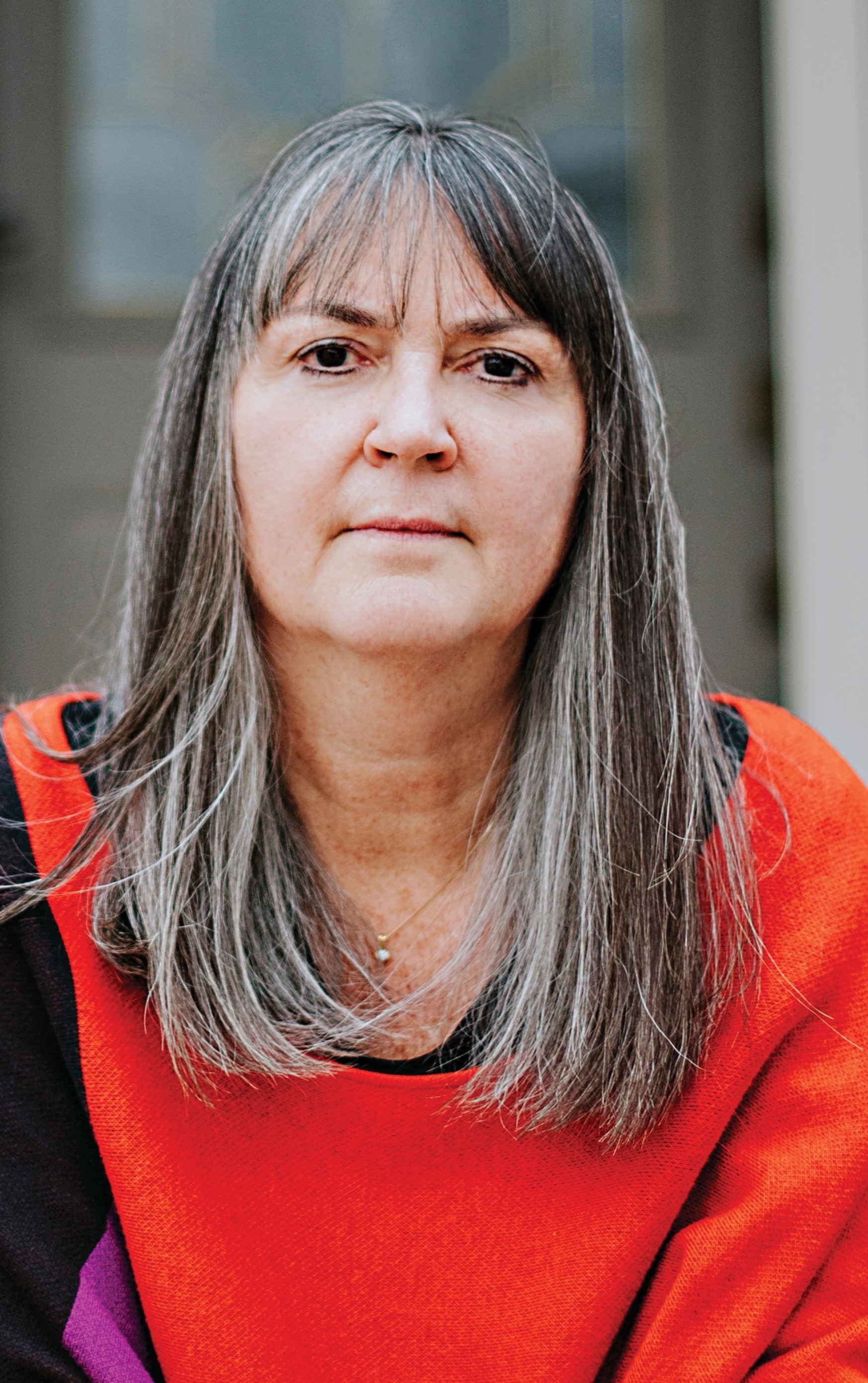 A person with long, gray and brown hair with bangs wears a red sweater and looks into the camera in front of a gray stone building.