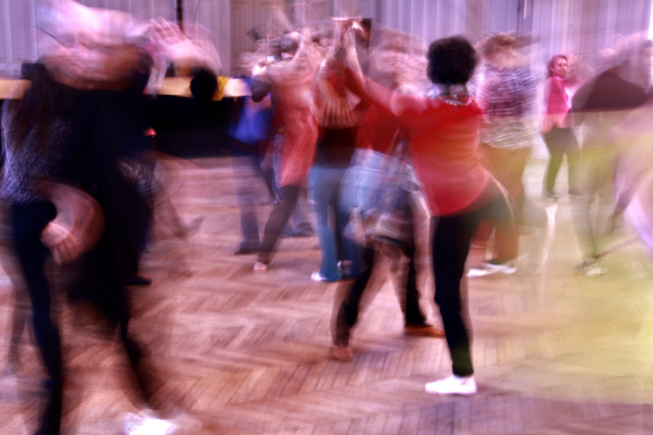 A group of people move energetically across a wooden floor, their bodies blurred by motion as they raise their arms during a dance or movement workshop.