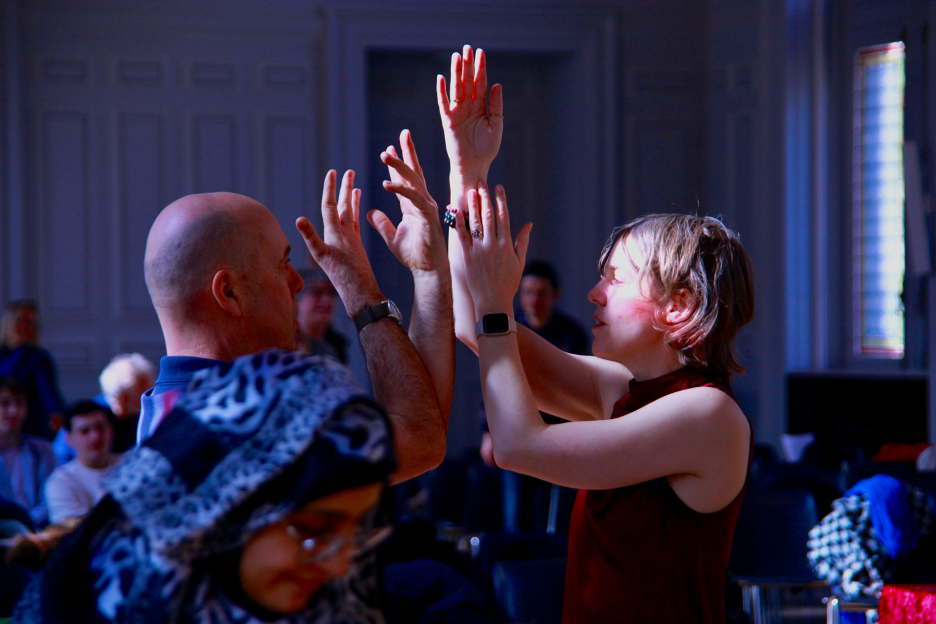 Two people face each other in a softly lit room, raising their hands toward one another as part of a guided movement or dance exercise, with seated participants blurred in the background.