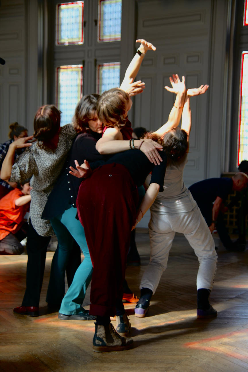 A group of dancers lean and reach together in a contemporary dance pose inside a large room with tall windows, their arms extended and bodies intertwined.