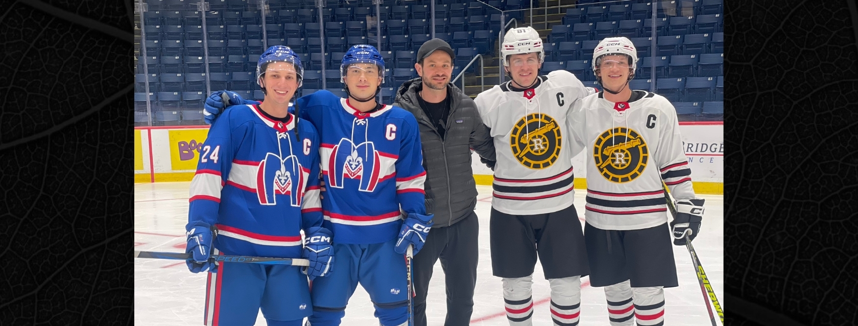 Two men stand on arena ice in blue hockey uniforms stand in a row with a man in a grey coat, black pants and black ball cap beside two men in white hockey uniforms smiling into the camera with arena stands behind them.