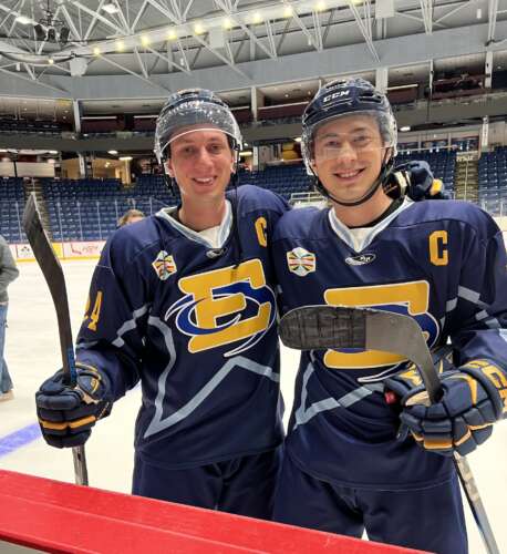 Two men in full hockey gear with blue and yellow jerseys stand smiling on the ice holding their hockey sticks.