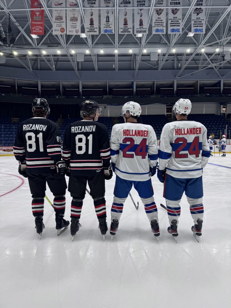A group of four hockey players, two in black and two in white, stand with their backs to the cameras to display their names and numbers on the ice holding hockey sticks.