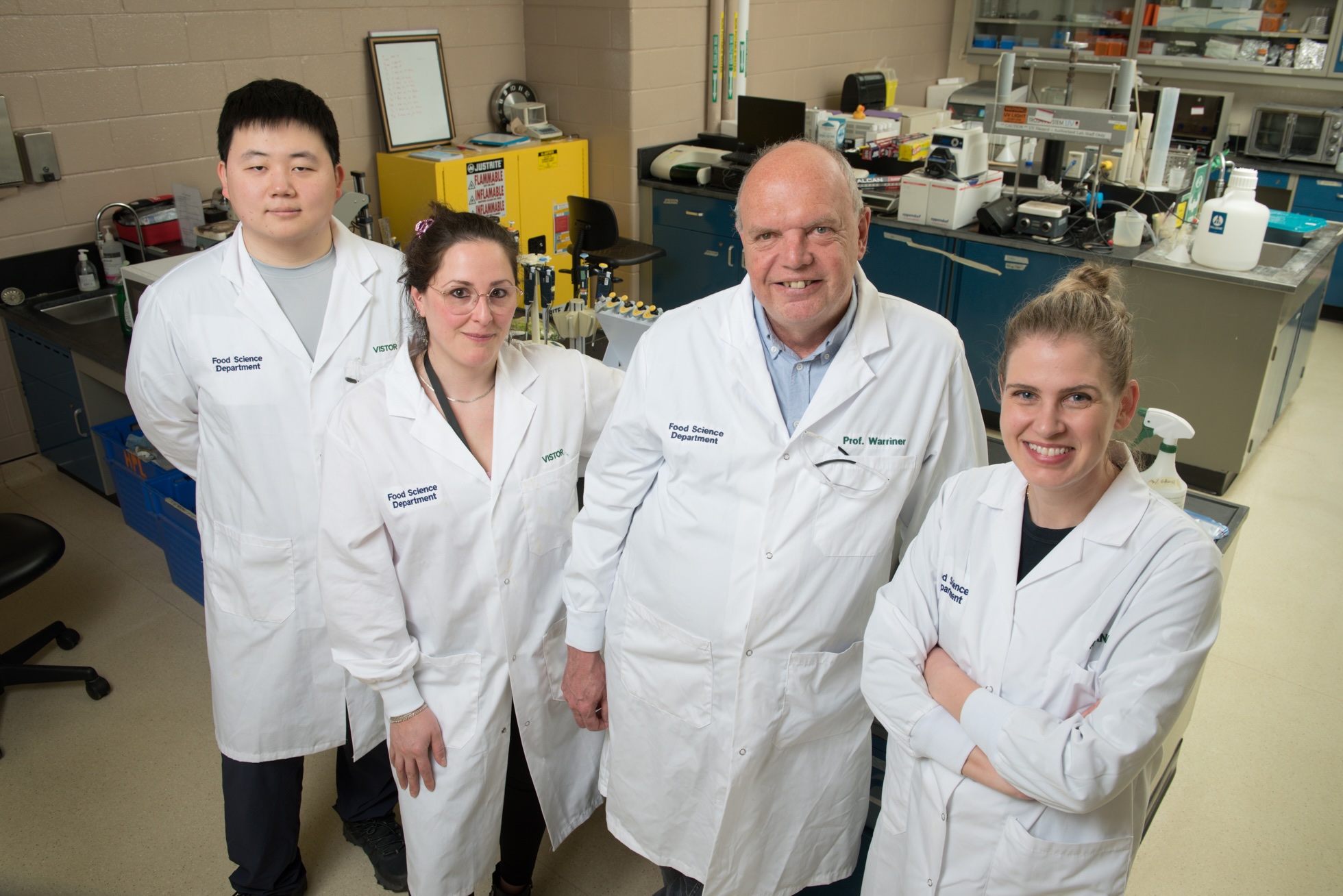 Four people in white lab coats stand together in a food science laboratory, surrounded by lab benches, bottles, and scientific equipment.