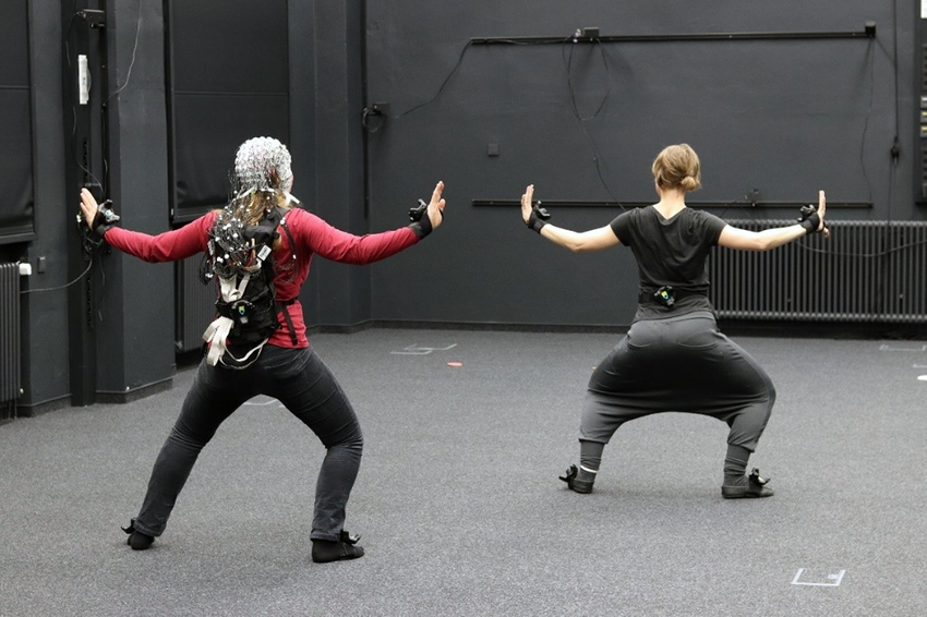 Two people wearing motion-capture sensors and headgear stand in a wide stance with arms raised in a laboratory space, participating in a movement research experiment