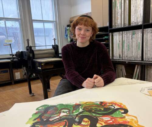 A person with red curly hair wearing a black headband and purple sweater sits on a stool leaning on a table with art prints smiling into the camera with a printing press, shelves and a window behind her.